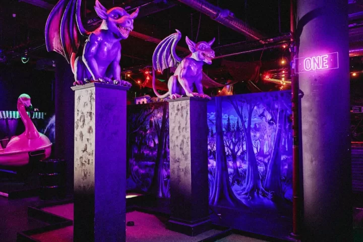 Two purple gargoyles on stone plinths under ultraviolet light beside a crazy golf hole