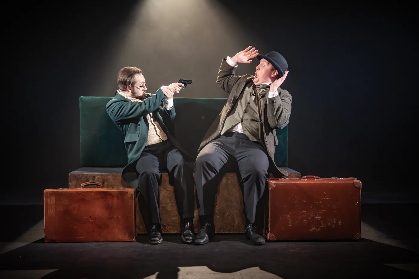 Two actors sit on a bench with suitcases, one aiming a revolver, the other shielding his eyes
