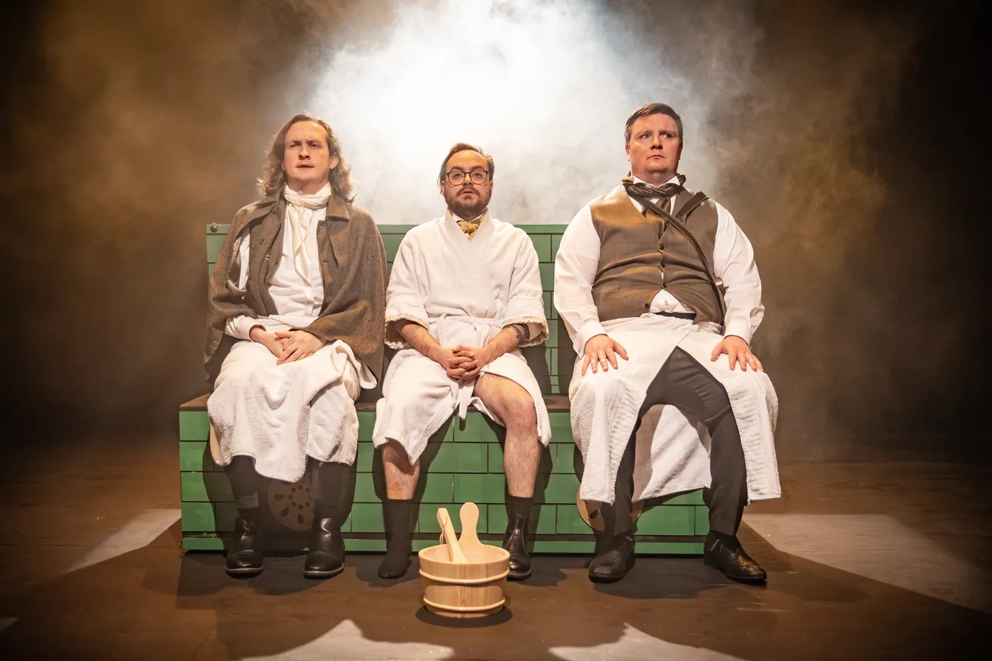 Three actors in Victorian nightshirts sit side by side on a green bench in smoky light